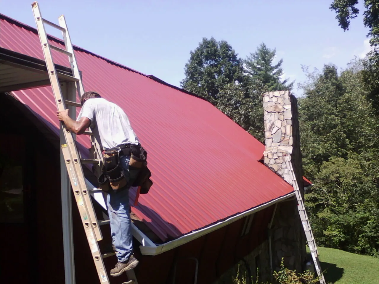Roofer on a ladder beside a red metal roof and stone chimney — Soffit & Fascia Repair by Roofing Brothers in Pleasant Dale